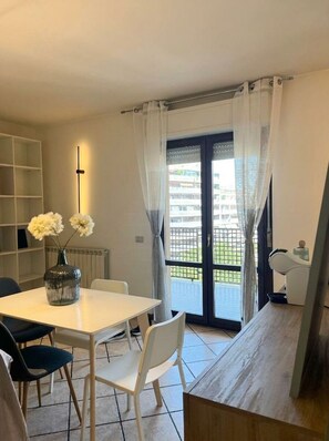 A dining area with a table and chairs, a vase with flowers, a radiator, and a view through a glass door to an outdoor balcony.