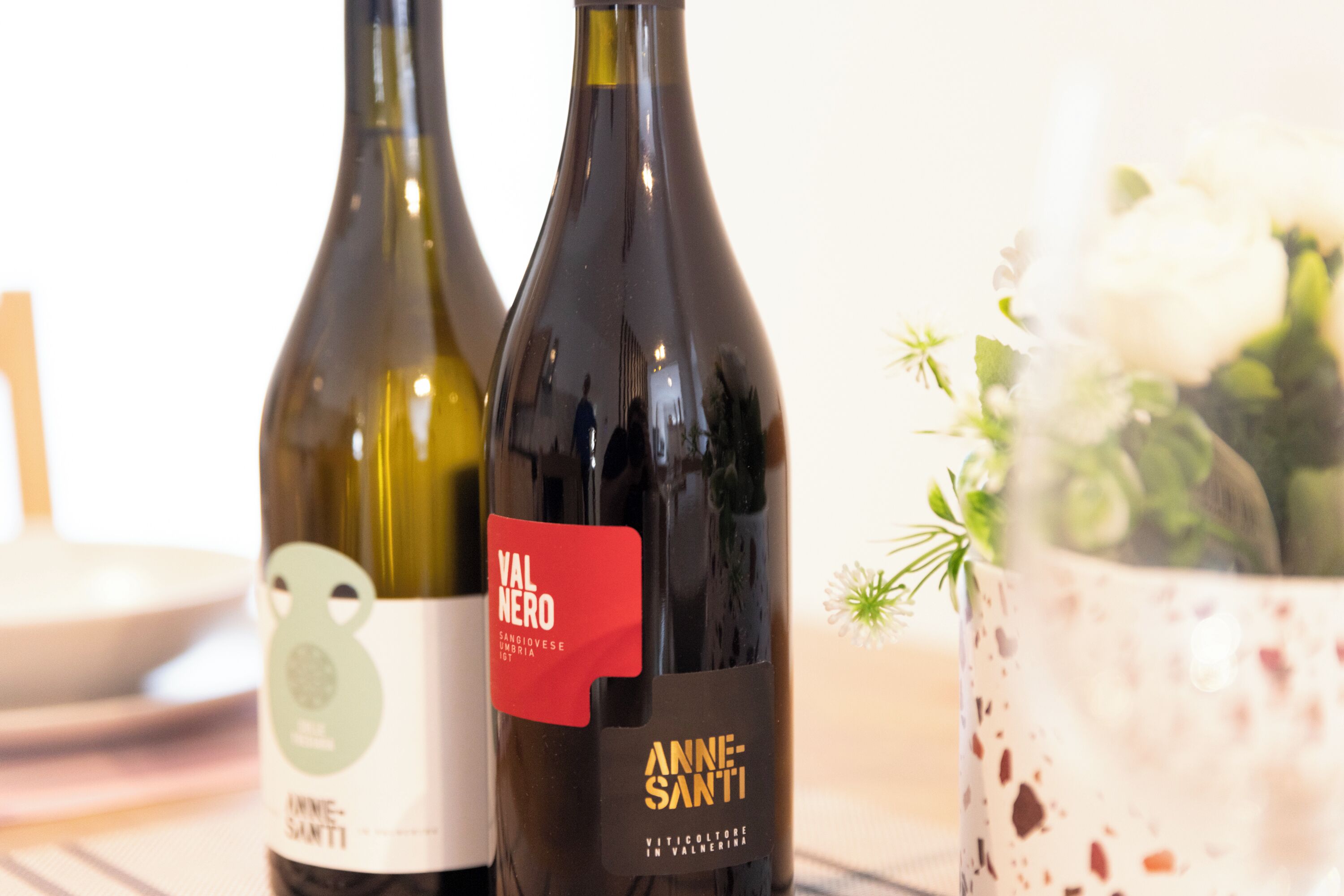Two wine bottles with labels, one labeled "Val Nero" and the other "Anne-Santi," placed on a table next to a bowl and a vase with flowers.