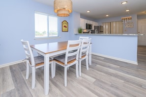 A modern kitchen with a central island, white cabinetry, and a dining area with a wooden table and white chairs. - Walk to the Beach 2 BR Condo with a Shared Pool! Colony III 201 (Surfside Beach)