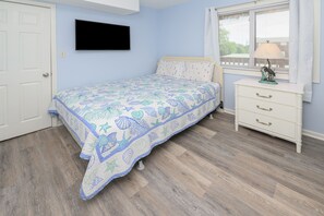 Ein Schlafzimmer mit Bett, Kommode, einem Fenster mit Jalousien und einem an der Wand befestigten Fernseher.