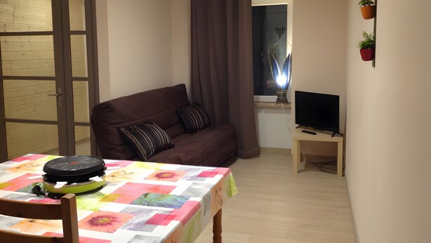 Ein Wohnzimmer mit einer Couch, einem Tisch mit einer Decke, einem Fernseher und einem Fenster mit Vorhängen.