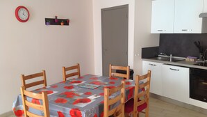 Ein Essbereich mit einem Tisch, bedeckt mit einer blumigen Tischdecke, Holzsesseln, einer Uhr an der Wand und einer Küchenzeile mit Schränken und einer Spüle.