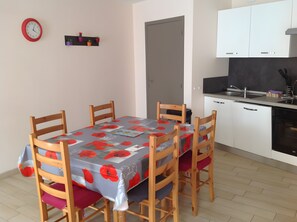 A dining area with a table covered in a floral tablecloth, wooden chairs, a clock on the wall, and a kitchenette with cabinets and a sink. - Apartment 'La Maison Des Bains De César' with Mountain View and Wi-Fi (Mont-Dore)