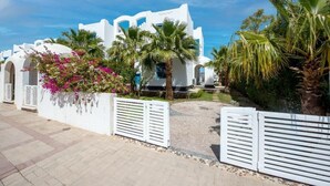 A white building with arched doorways, surrounded by palm trees and pink flowers.