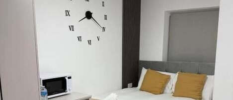 Habitación de hotel con cama, mesita de noche con microondas, refrigerador pequeño y un reloj de pared.