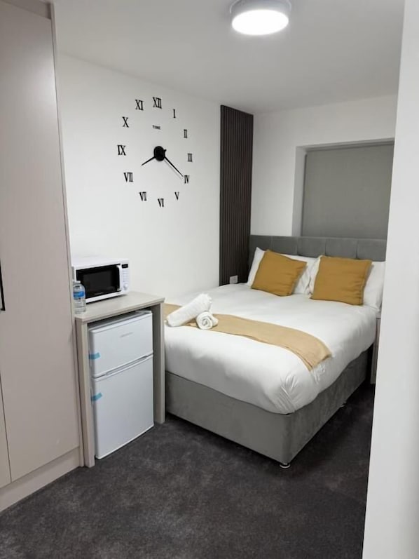 A hotel room with a bed, a bedside table with a microwave, a small refrigerator, and a wall clock.