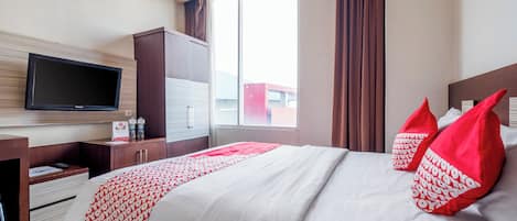 Ett hotellrum med en säng, en tv som är monterad på väggen, ett skrivbord med lådor och ett fönster med gardiner.