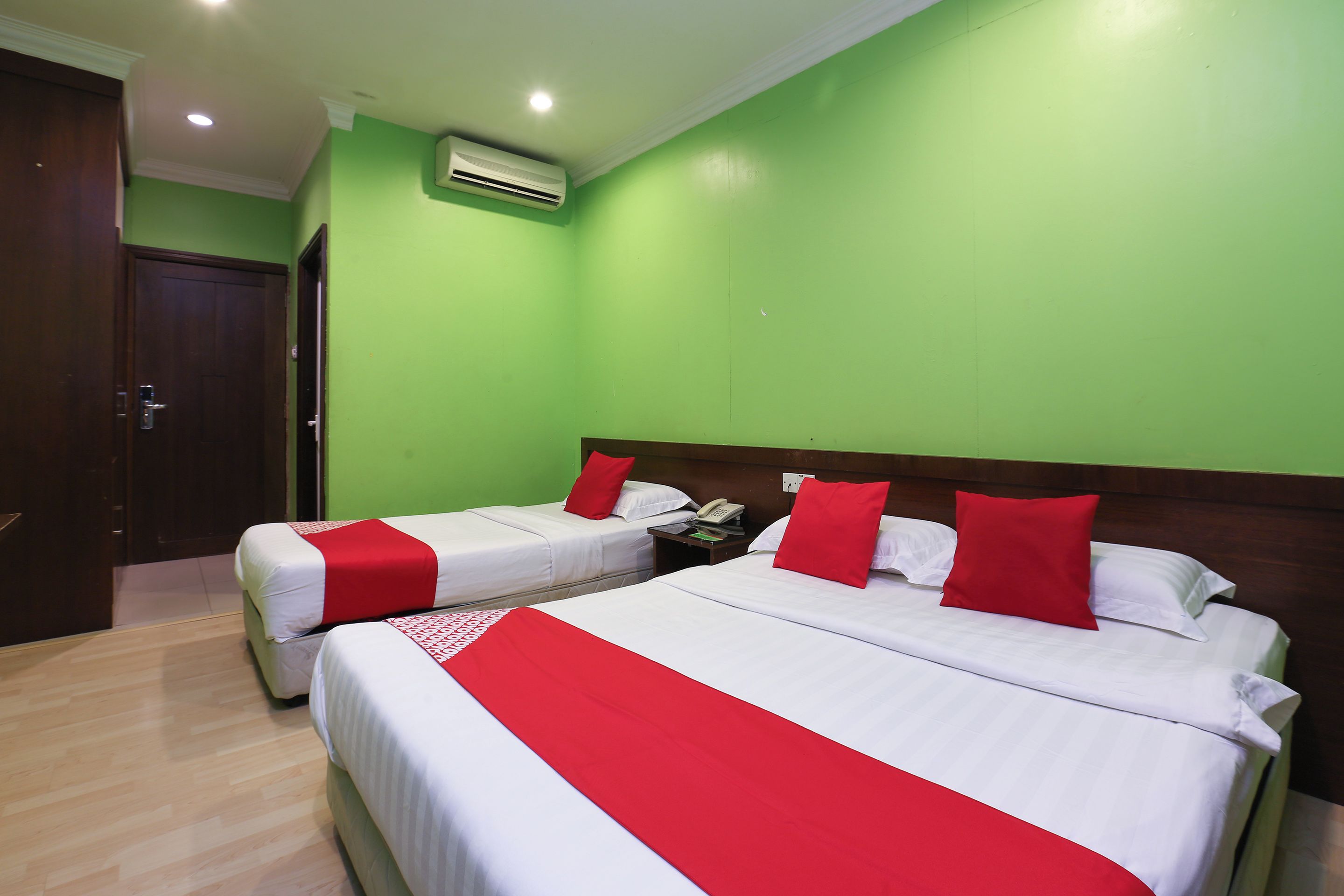 A hotel room with two beds, each with white bedding and red accents, a wooden headboard, a nightstand with a phone, and a green wall.