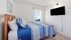 Un dormitorio con cama, televisión montada en la pared, ventana con persianas y puerta.