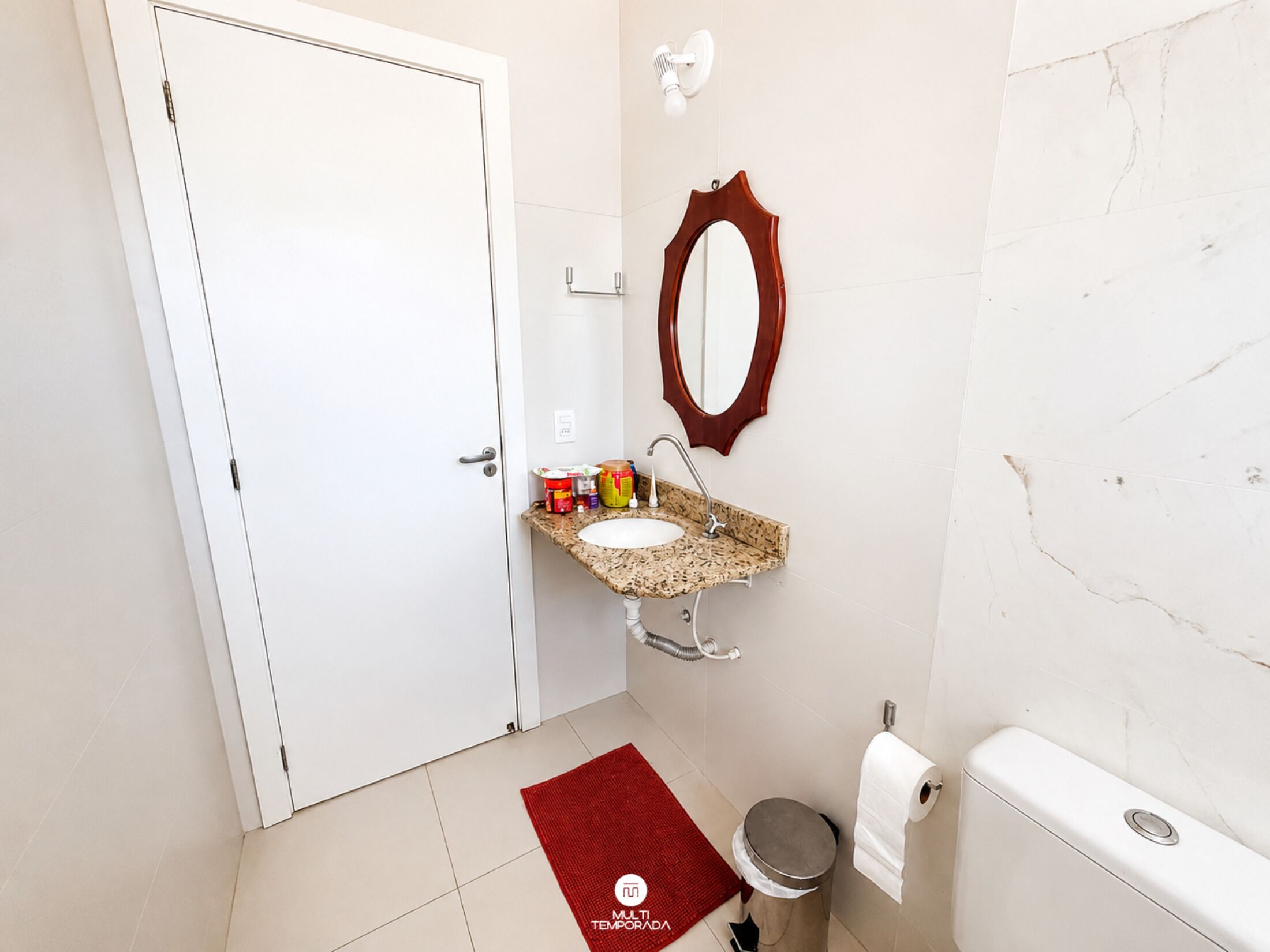 Ein Badezimmer mit einer weißen Tür, einem Waschbecken mit Granitplatte, einem Spiegel, einer Toilette und einem roten Teppich.
