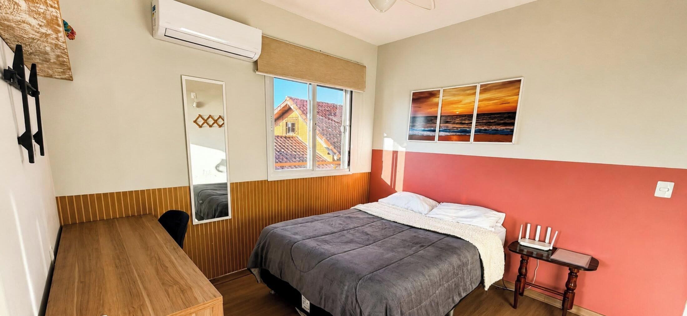A bedroom with a bed, a desk, a chair, a window with a view of a house, and an air conditioning unit.