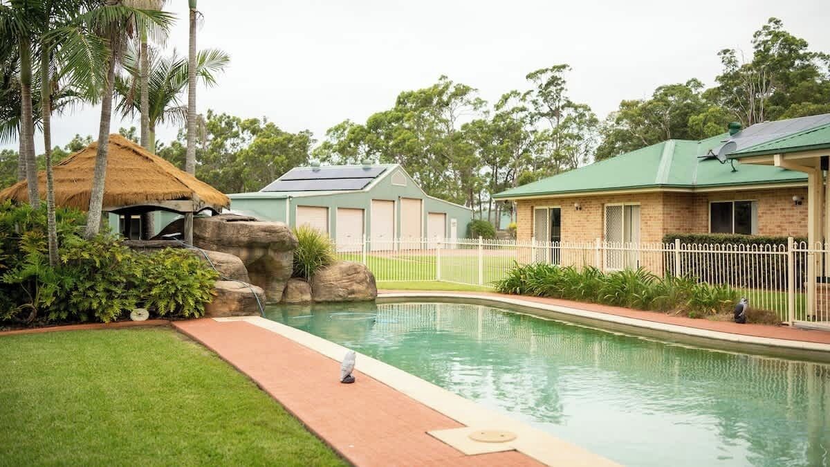 A swimming pool with a brick building, a thatched-roof structure, and a fenced area.
