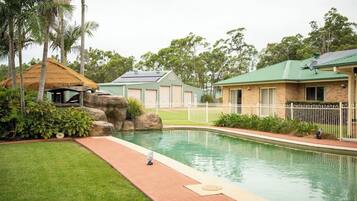 A swimming pool with a brick building, a thatched-roof structure, and a fenced area.
