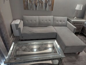 Una sala de estar moderna con un sofá seccional gris, una mesa de centro de vidrio y una mesita de noche con una lámpara.