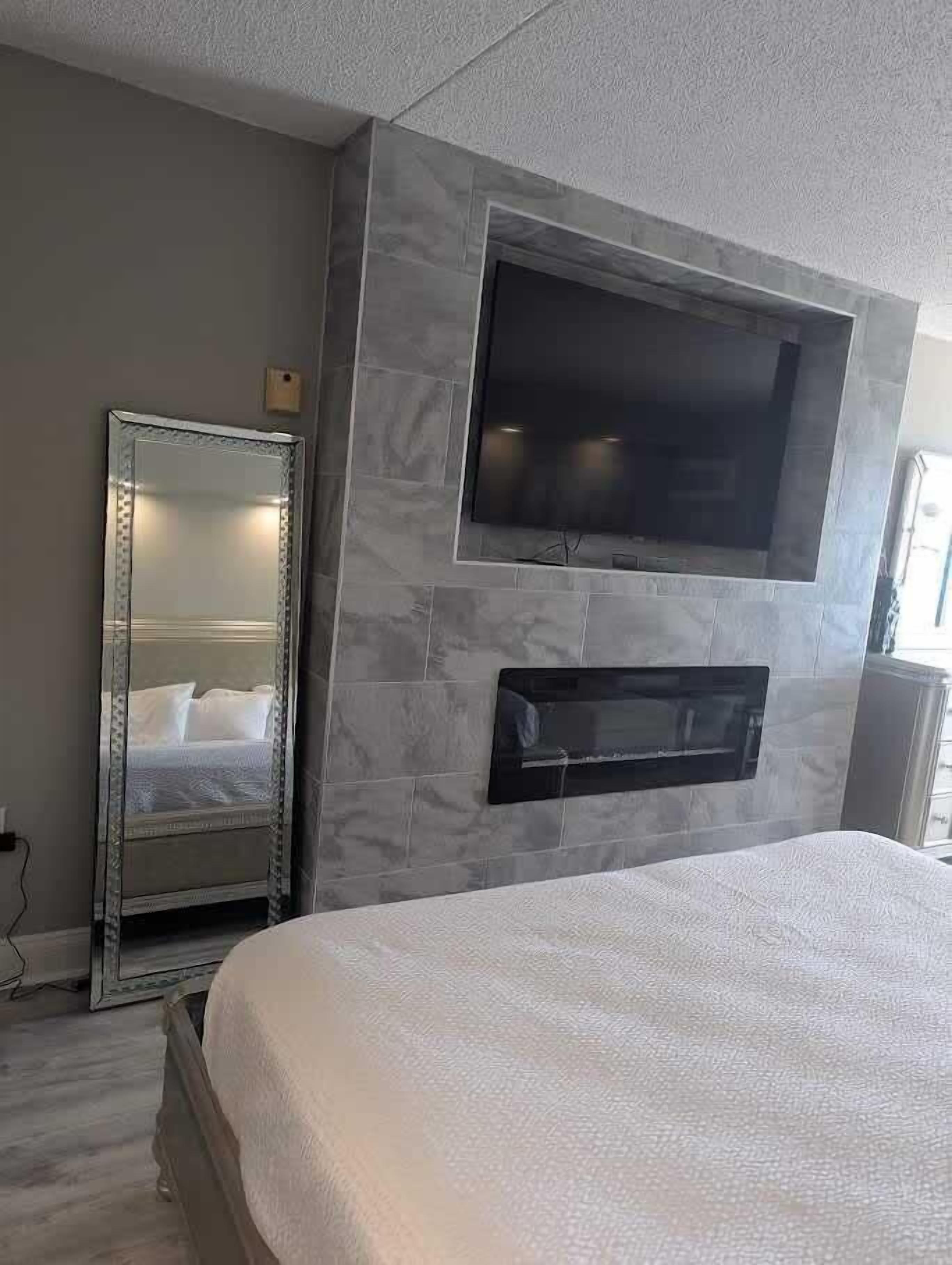 A bedroom with a bed, a large mirror, a flat-screen TV mounted on the wall, and a fireplace below the TV.