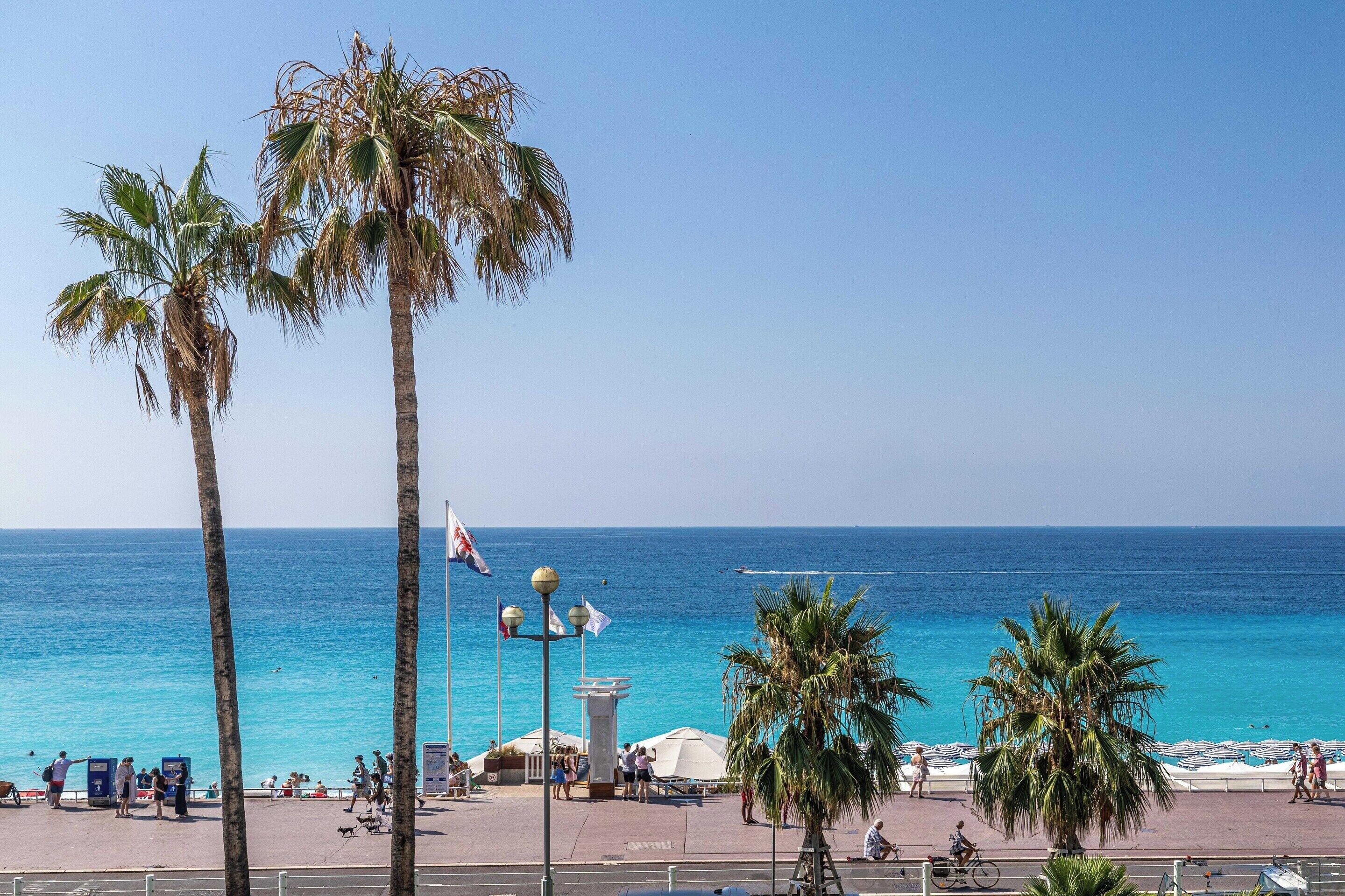 Un paseo marítimo con palmeras, un mar azul cristalino y algunas personas caminando.
