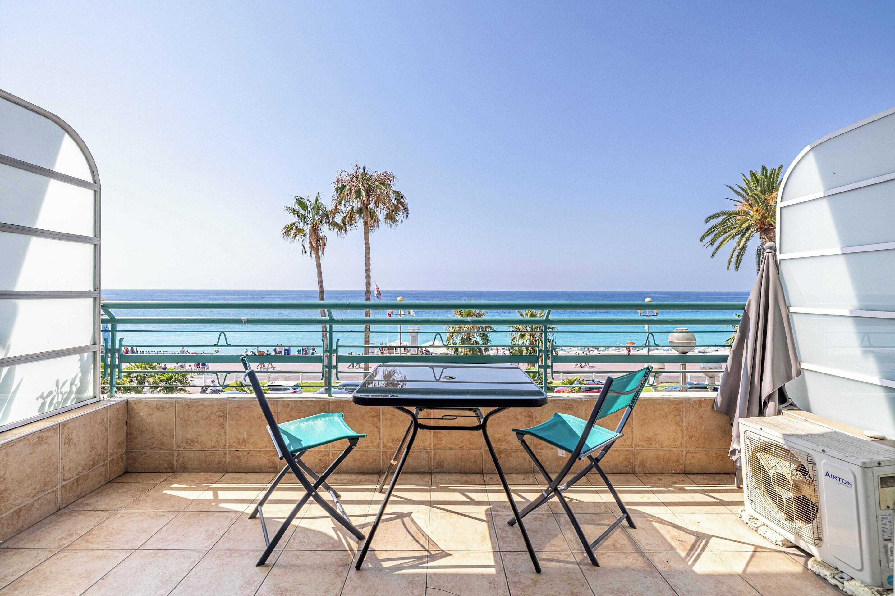 Un balcón con mesa y sillas, con vista a una playa con palmeras y cielo despejado.