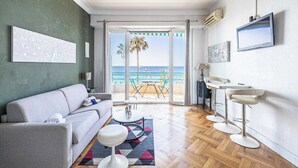 A modern living room with a sofa, a coffee table, and a view of the beach through the glass door. - Charming Apartment a/c 2P by the Seafront - Nice (Nice)