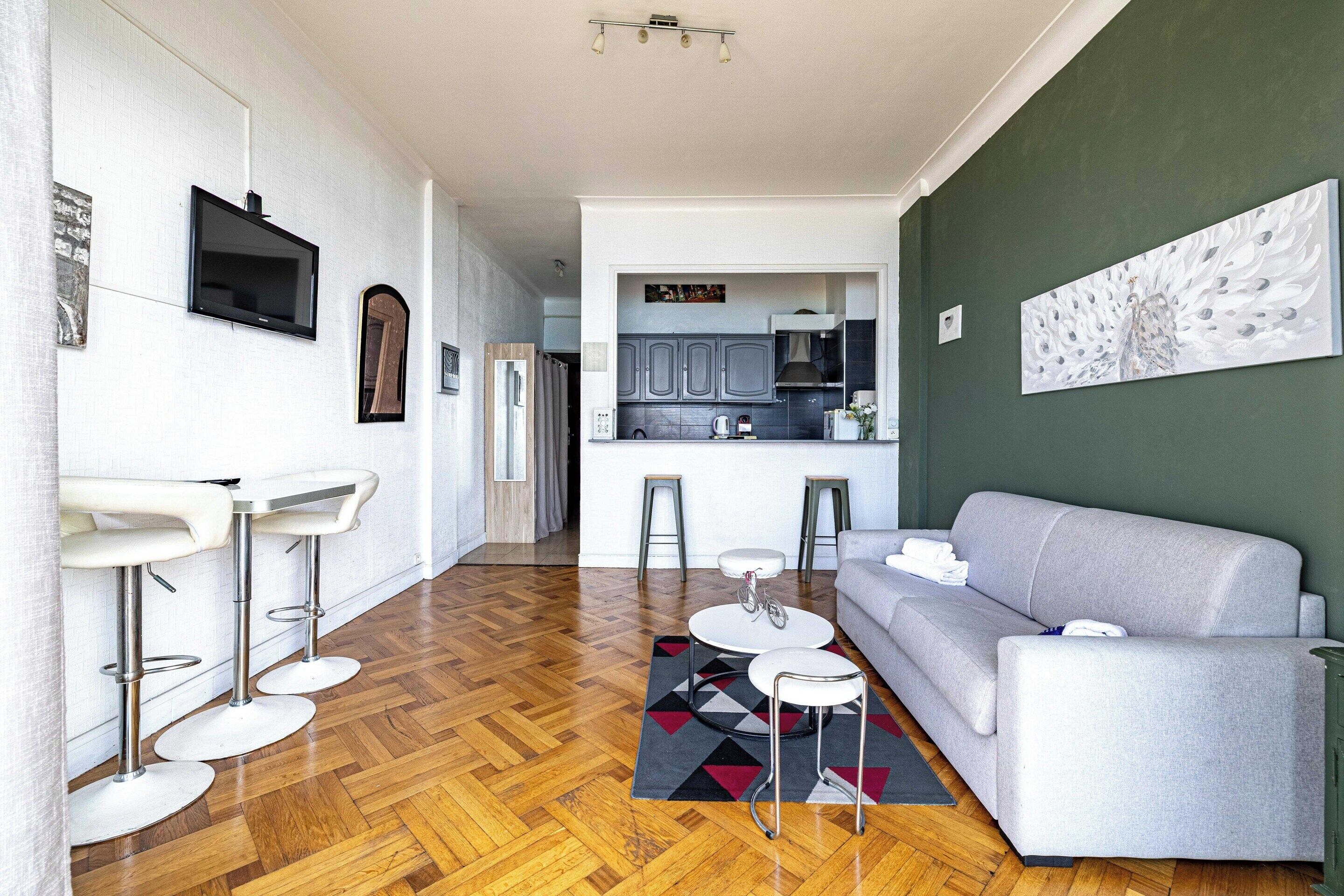 Una sala de estar moderna con un sofá gris, piso de madera en espiga y una cocina al fondo.