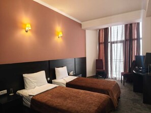 Una habitación de hotel con dos camas, un escritorio con una silla, un televisor y una ventana con cortinas.