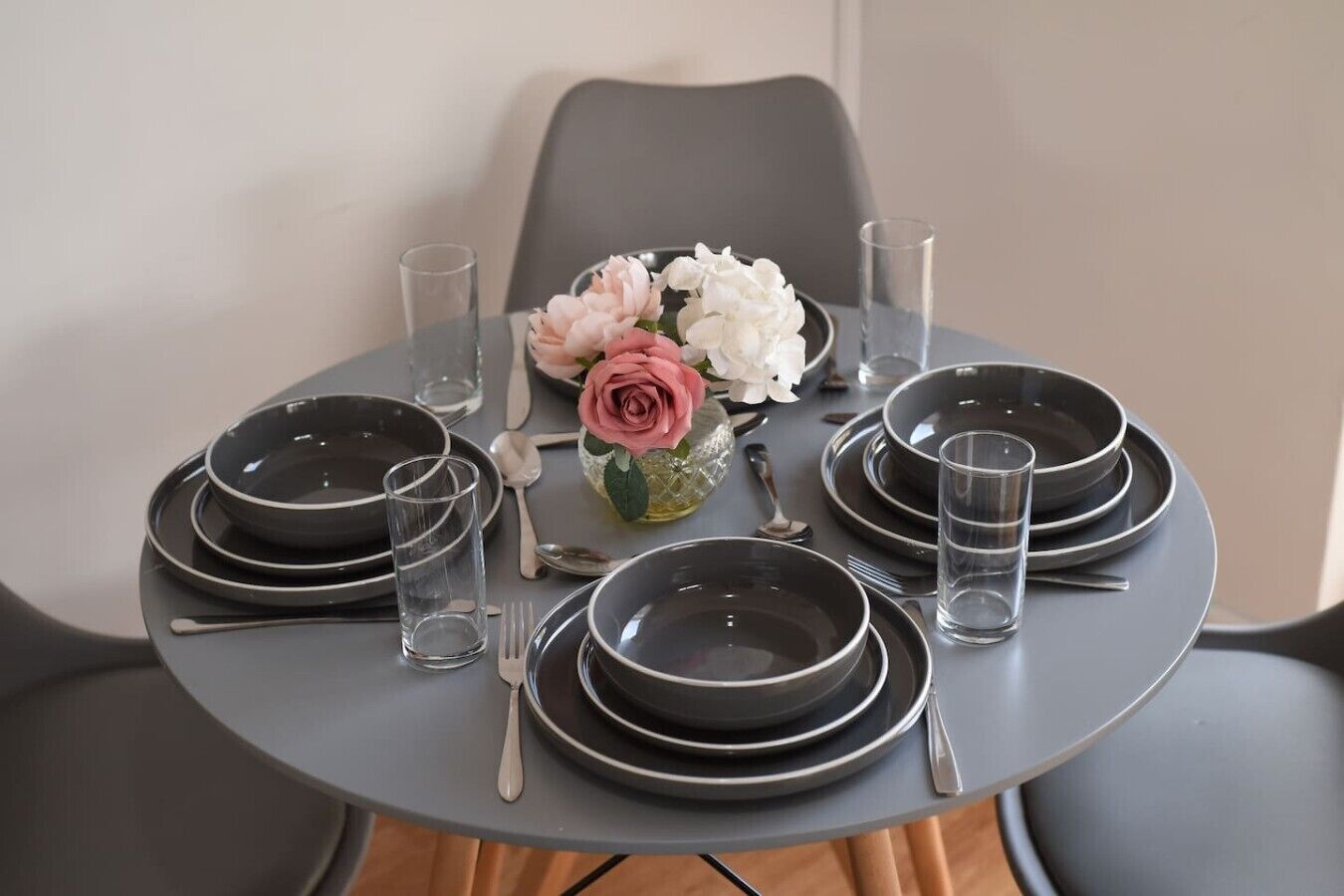 Ett runt bord dukat för fyra personer, med svarta tallrikar, klara glas och silverbestick. En vas med rosa och vita blommor står i mitten av bordet.