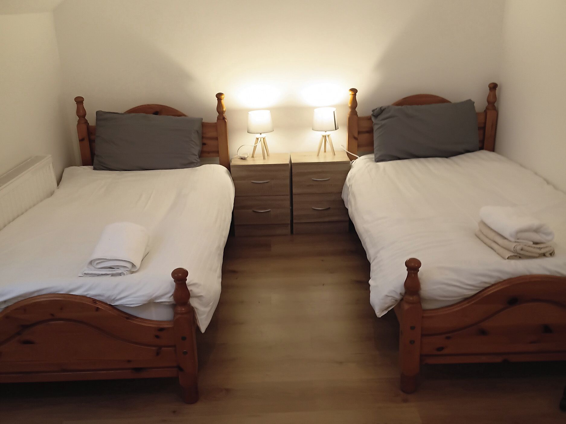 A twin room with two wooden beds, each with a pillow and a folded towel. There are bedside tables with lamps and a small mirror.