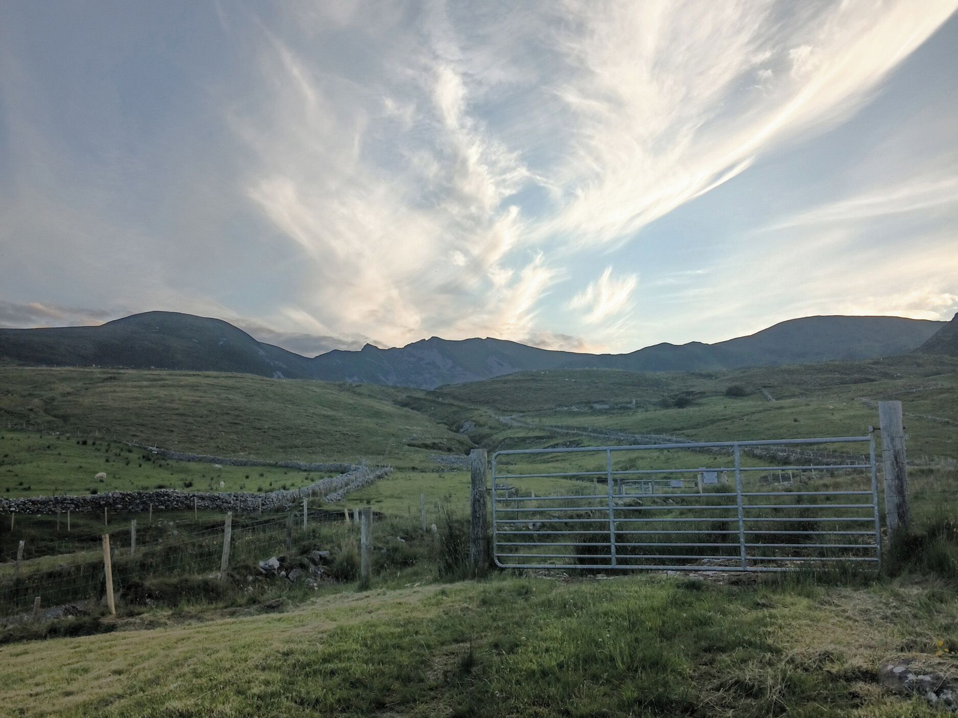 A rural landscape with a metal gate, stone wall, and grassy fields.