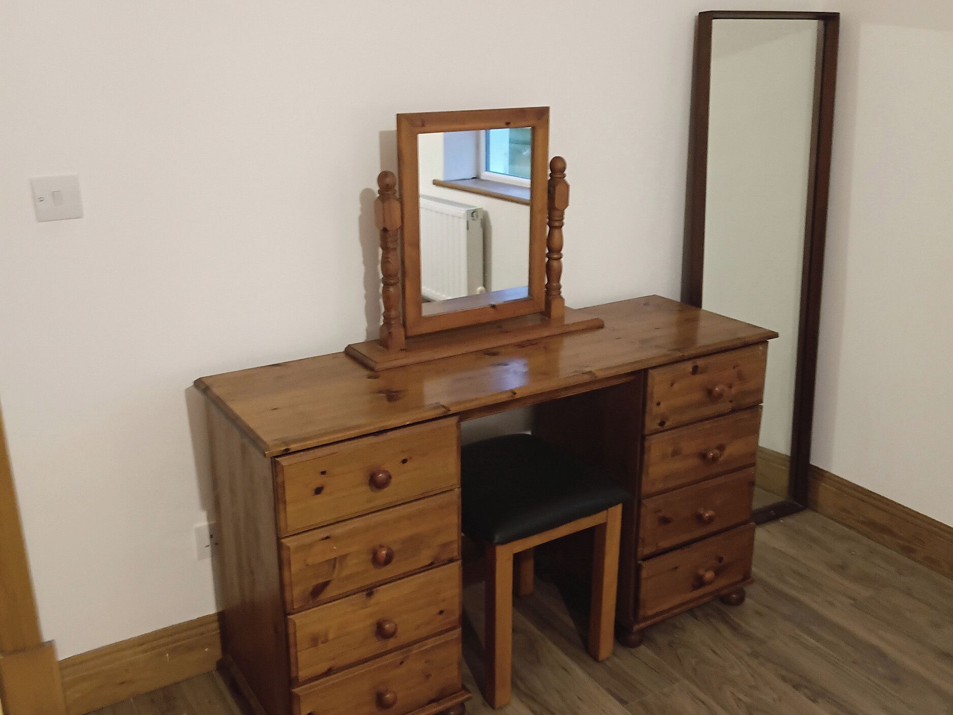 A wooden dresser with a mirror and a chair, a light switch on the wall, and a radiator in the reflection.
