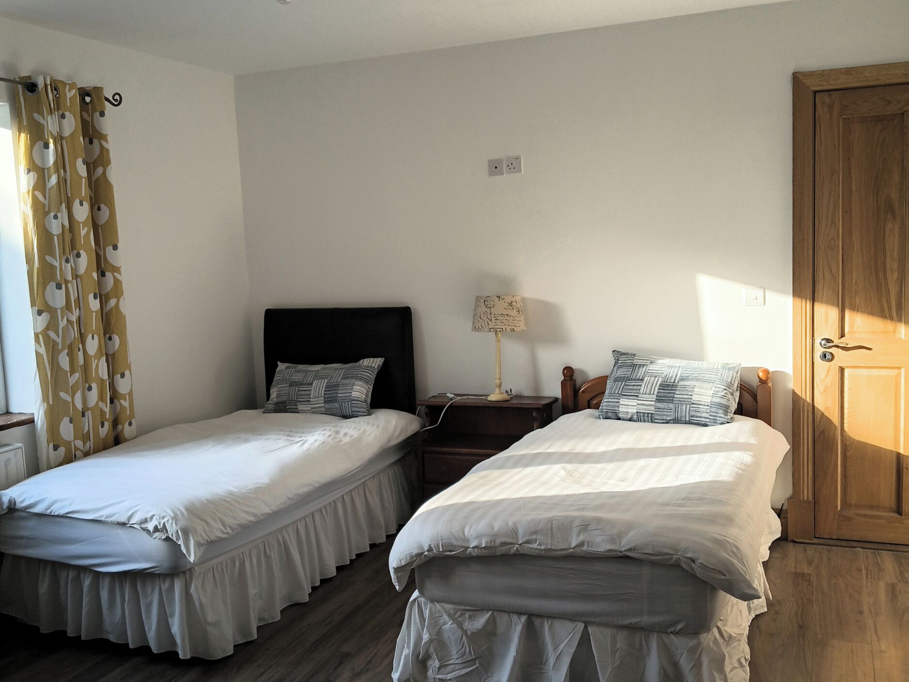 A bedroom with two single beds, a nightstand with a lamp, a wooden door, and a window with curtains.