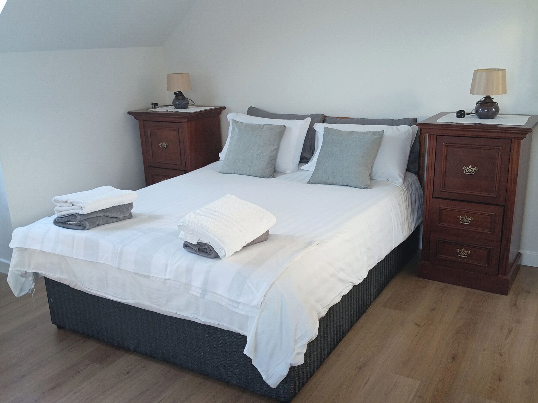 A neatly made bed with white linens, grey pillows, and a grey blanket. There are two bedside tables with lamps and a small drawer. The room has wooden flooring and white walls.
