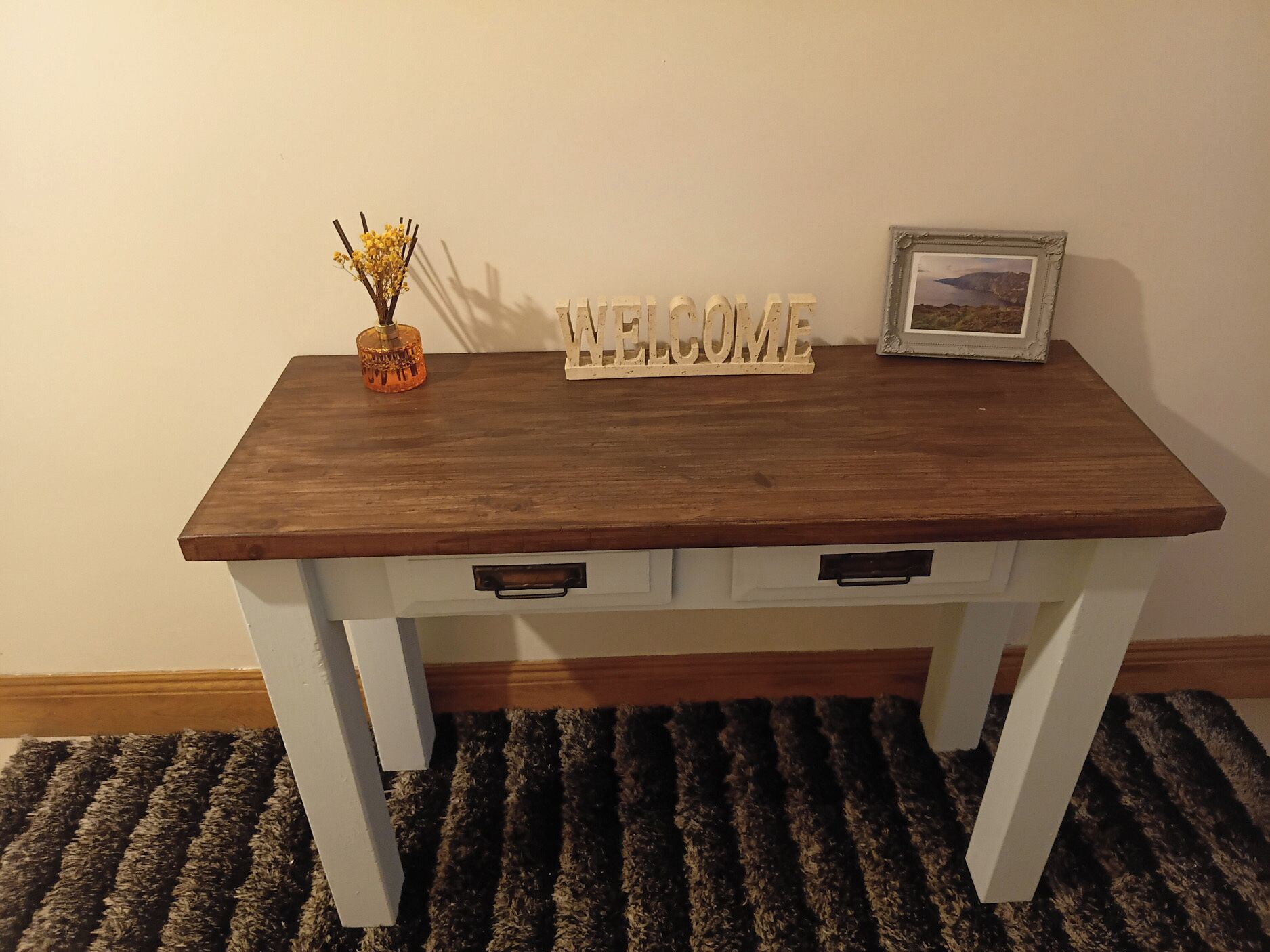 A wooden table with a 'WELCOME' sign, a small vase with flowers, and a framed picture.