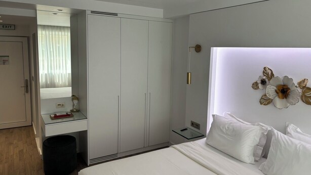 A hotel room with a bed, a bedside table, a mirror, a wardrobe, and a wall-mounted light fixture. - WHITE ROSE ATHENS RESIDENCE (Athens)