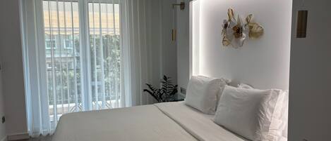 Un dormitorio moderno con una cama grande, una mesita de noche de vidrio y vistas a un balcón a través de cortinas traslúcidas.