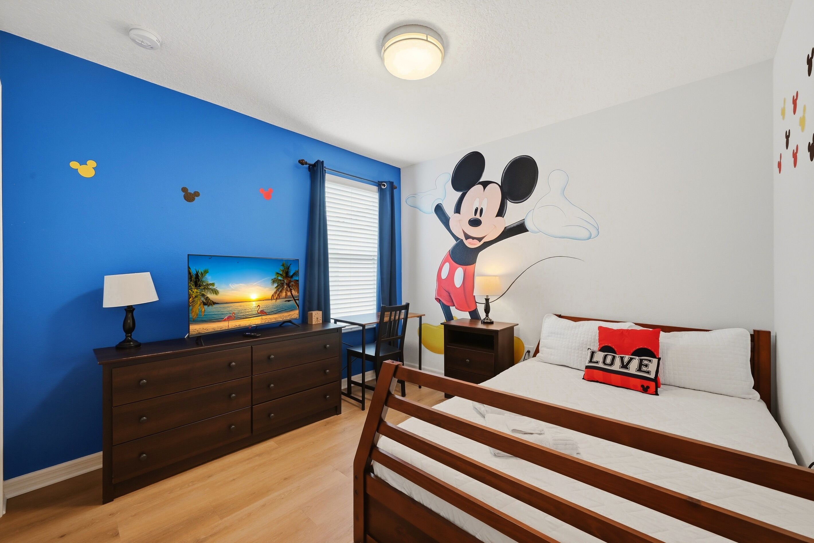 Une chambre avec une fresque murale représentant Mickey Mouse, un lit avec un coussin sur lequel est écrit le mot « AMOUR », un bureau avec une lampe et une table de chevet avec une lampe.
