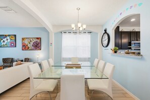 Uma sala de jantar moderna com uma mesa de vidro, cadeiras brancas, um lustre e um relógio na parede.