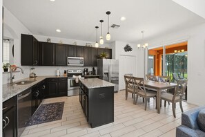 A modern kitchen with dark cabinets, a central island, and a dining area with a table and chairs. - Modern 6BRPoolVilla w/HotTub &GameRoom Near Disney (Kissimmee)