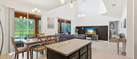 A spacious kitchen with a central island, wooden dining table, and chairs, with a view of a swimming pool through large windows.