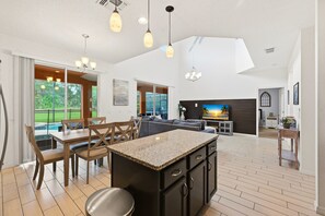 A spacious kitchen with a central island, wooden dining table, and chairs, with a view of a swimming pool through large windows. - Modern 6BRPoolVilla w/HotTub &GameRoom Near Disney (Kissimmee)