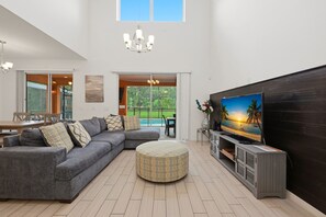 A spacious living area with a sectional sofa, a round ottoman, a flat-screen TV, and a dining area with a table and chairs. - Modern 6BRPoolVilla w/HotTub &GameRoom Near Disney (Kissimmee)