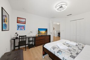 Ein Schlafzimmer mit einem Bett, einem Schreibtisch mit Globus und Lampe, einem an der Wand hängenden Fernseher und einer Tür zu einem weiteren Raum.