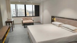 Ein Hotelzimmer mit einem großen Bett, einem kleinen Tisch, einem Sessel und einem Fenster mit Blick auf Gebäude.