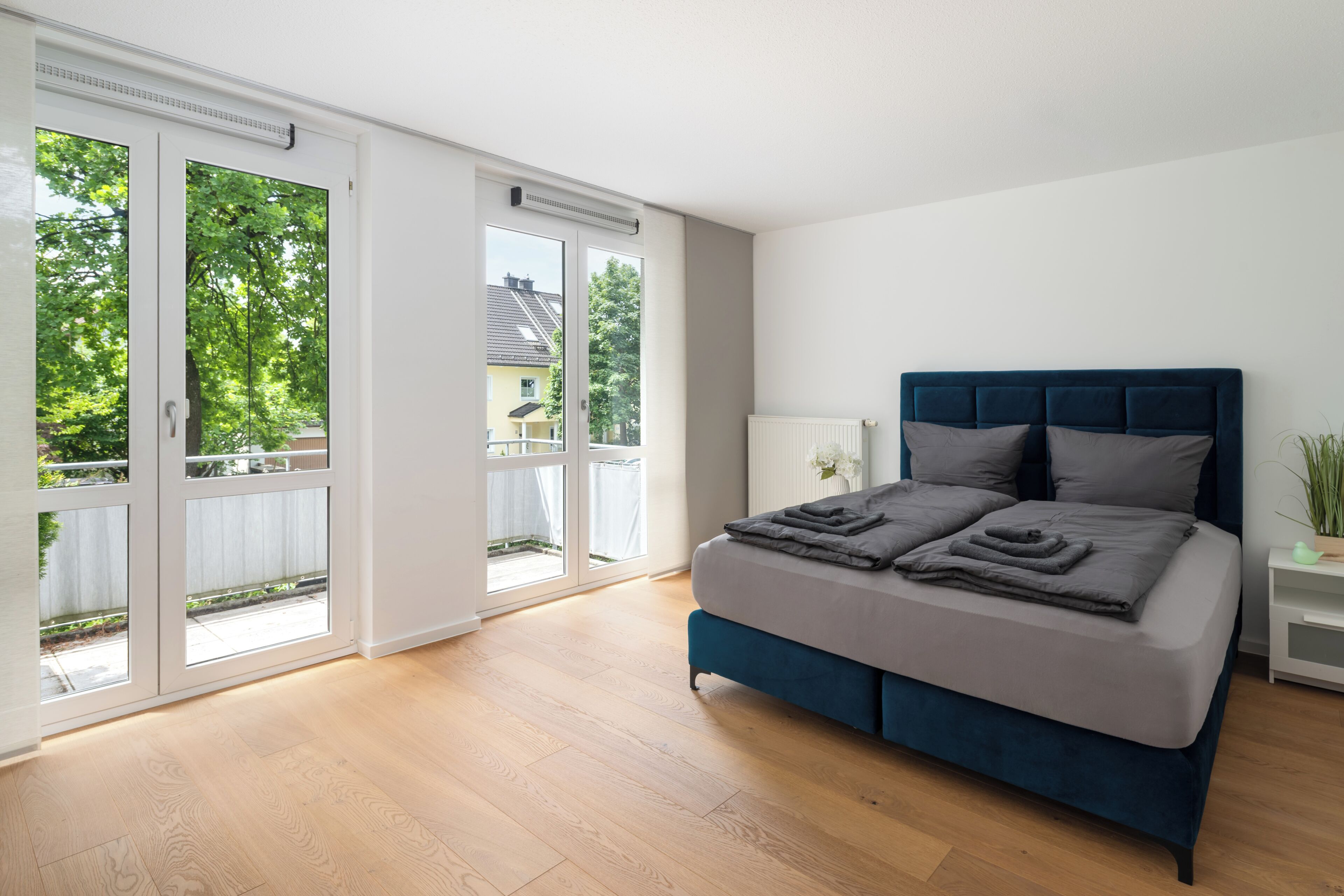 A bedroom with a bed, a nightstand, and a window with a view of greenery and buildings.