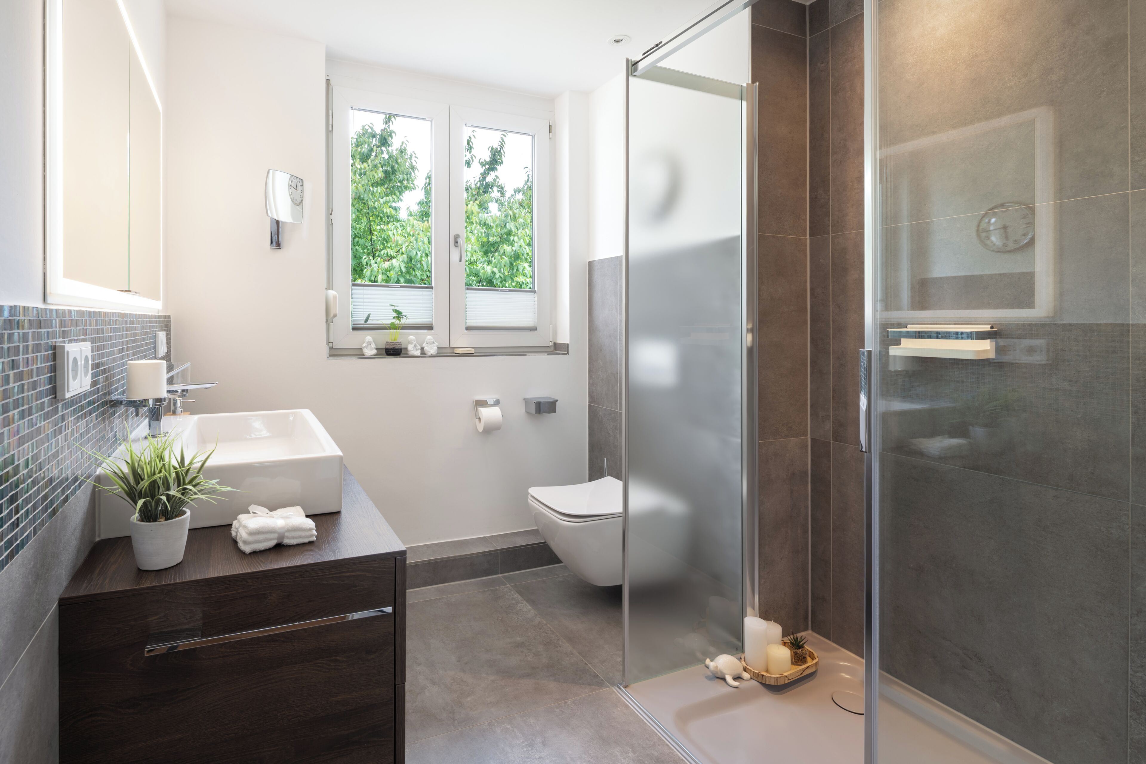 Une salle de bain moderne avec une douche à l’italienne en verre, un lavabo blanc et une fenêtre donnant sur un espace verdoyant.