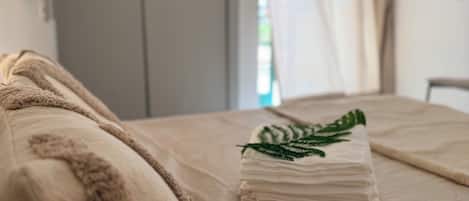 A bed with a beige blanket and pillows, a folded towel with a green leaf design, and a grey wardrobe in the background.