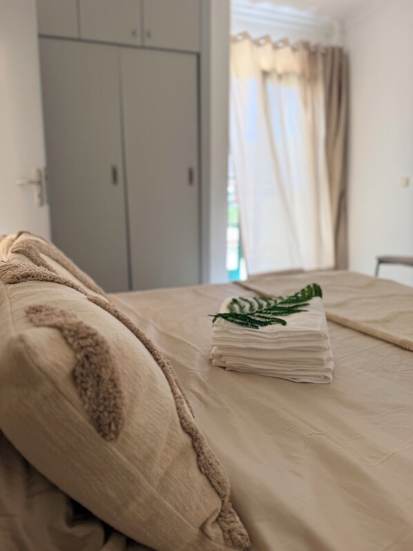 A bed with a beige blanket and pillows, a folded towel with a green leaf design, and a grey wardrobe in the background.