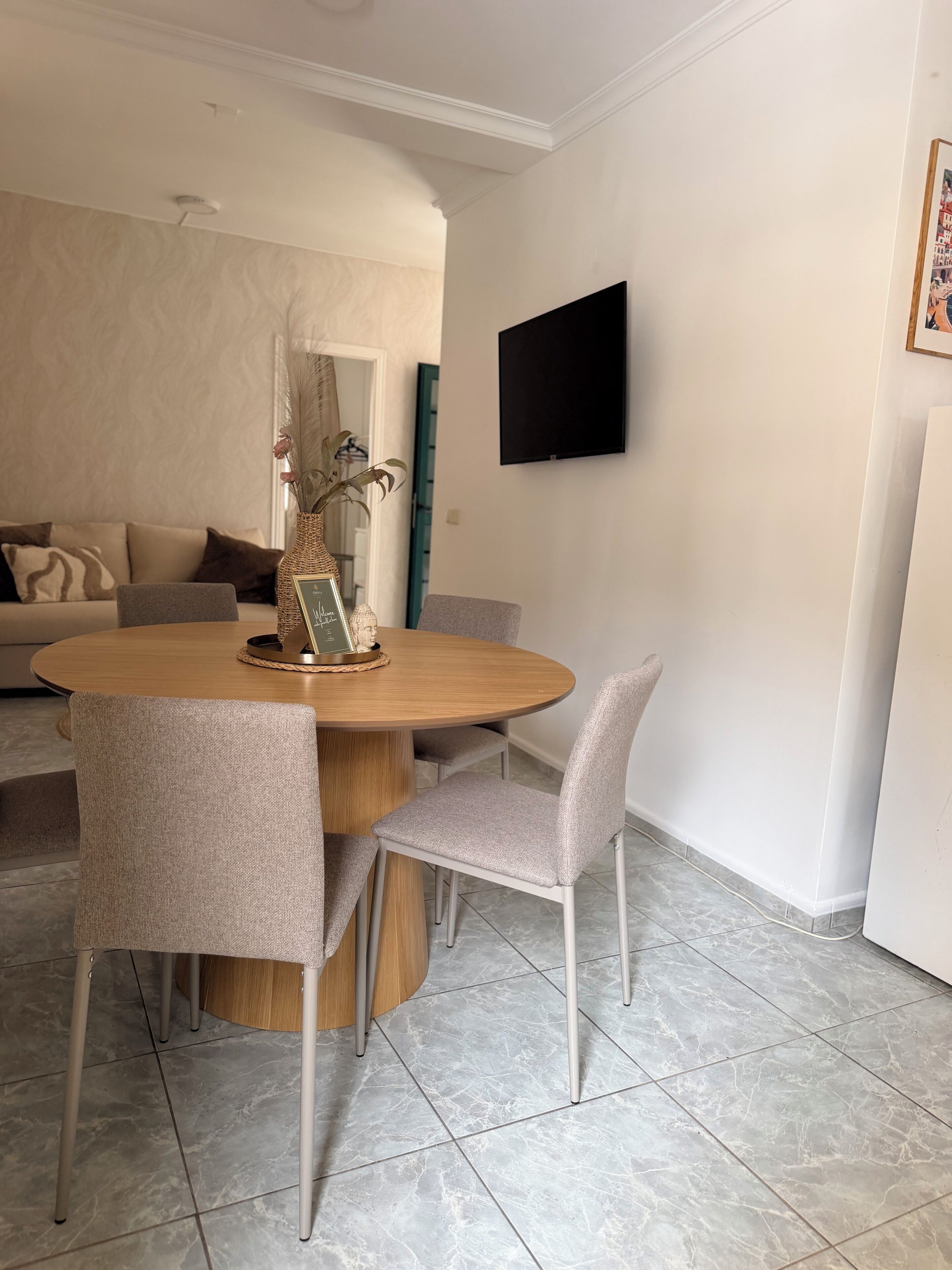 Un comedor con una mesa redonda de madera, cuatro sillas tapizadas de color gris, un jarrón decorativo y un televisor de pantalla plana montado en la pared.