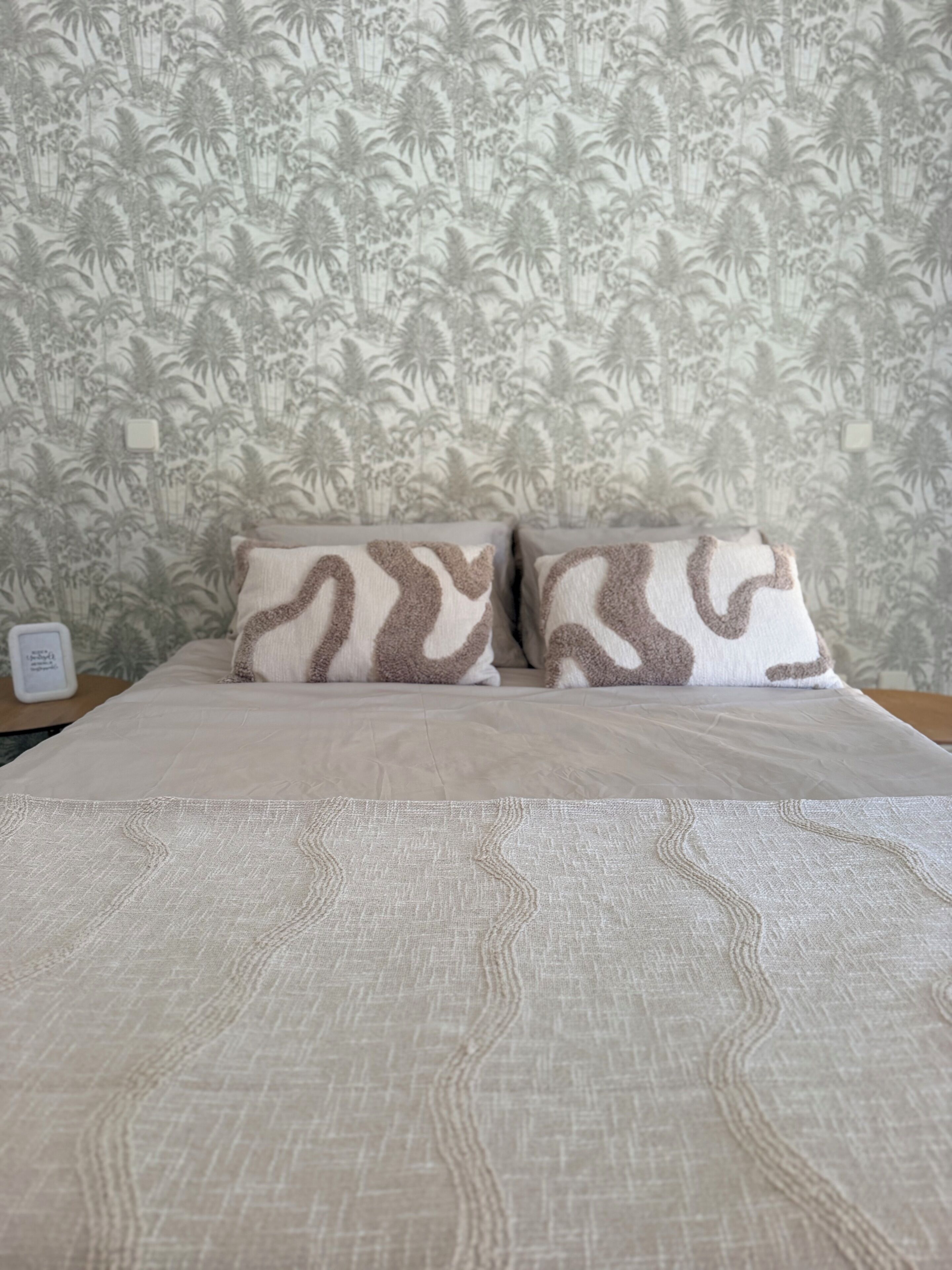 Una cama con colcha estampada y dos almohadas de diseño similar, frente a una pared con papel tapiz tropical.
