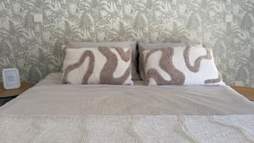 Una cama con colcha estampada y dos almohadas de diseño similar, frente a una pared con papel tapiz tropical.