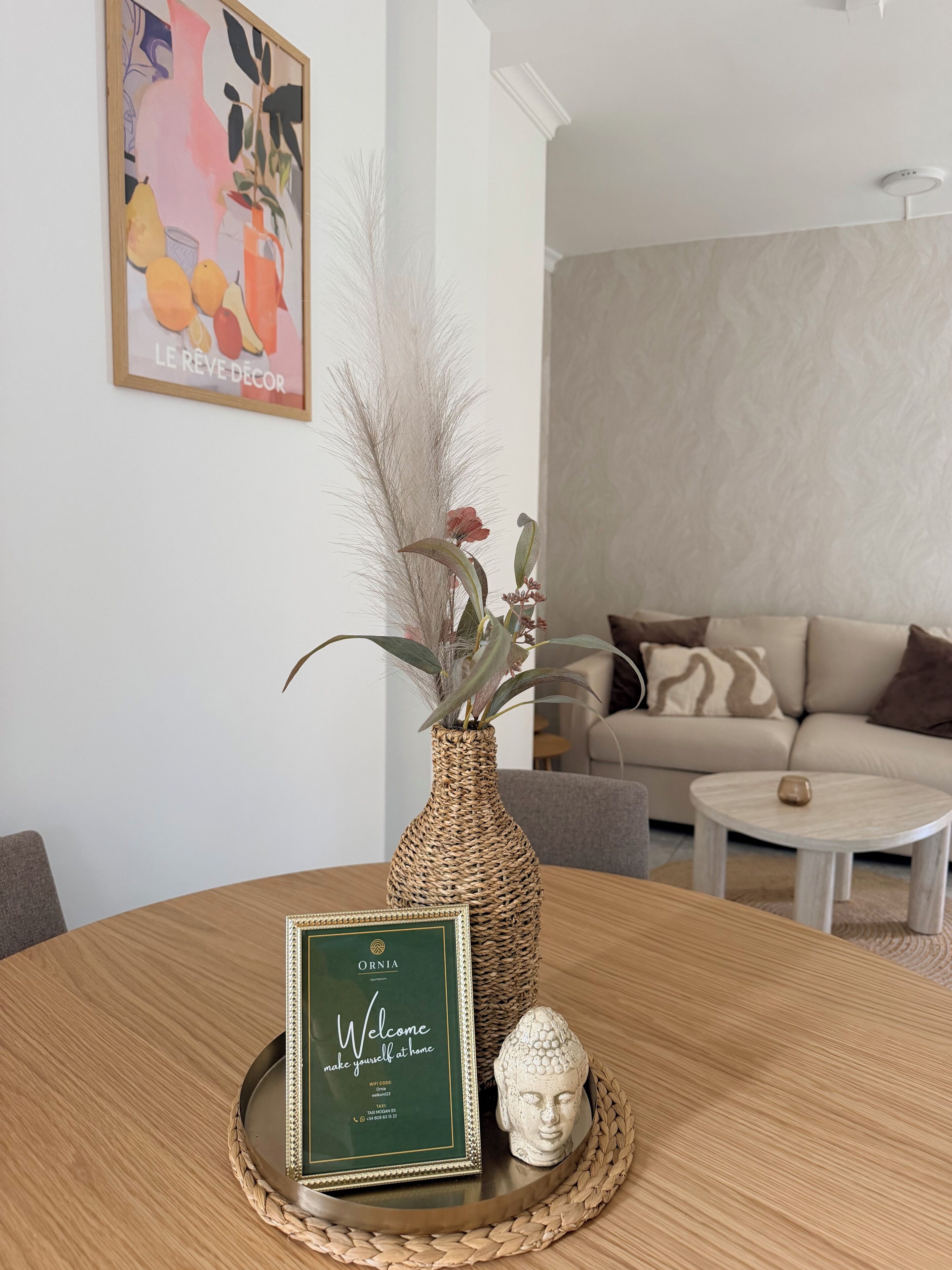 Una mesa de comedor con un cartel de bienvenida enmarco, un jarrón con plantas secas y una escultura de cabeza de Buda.