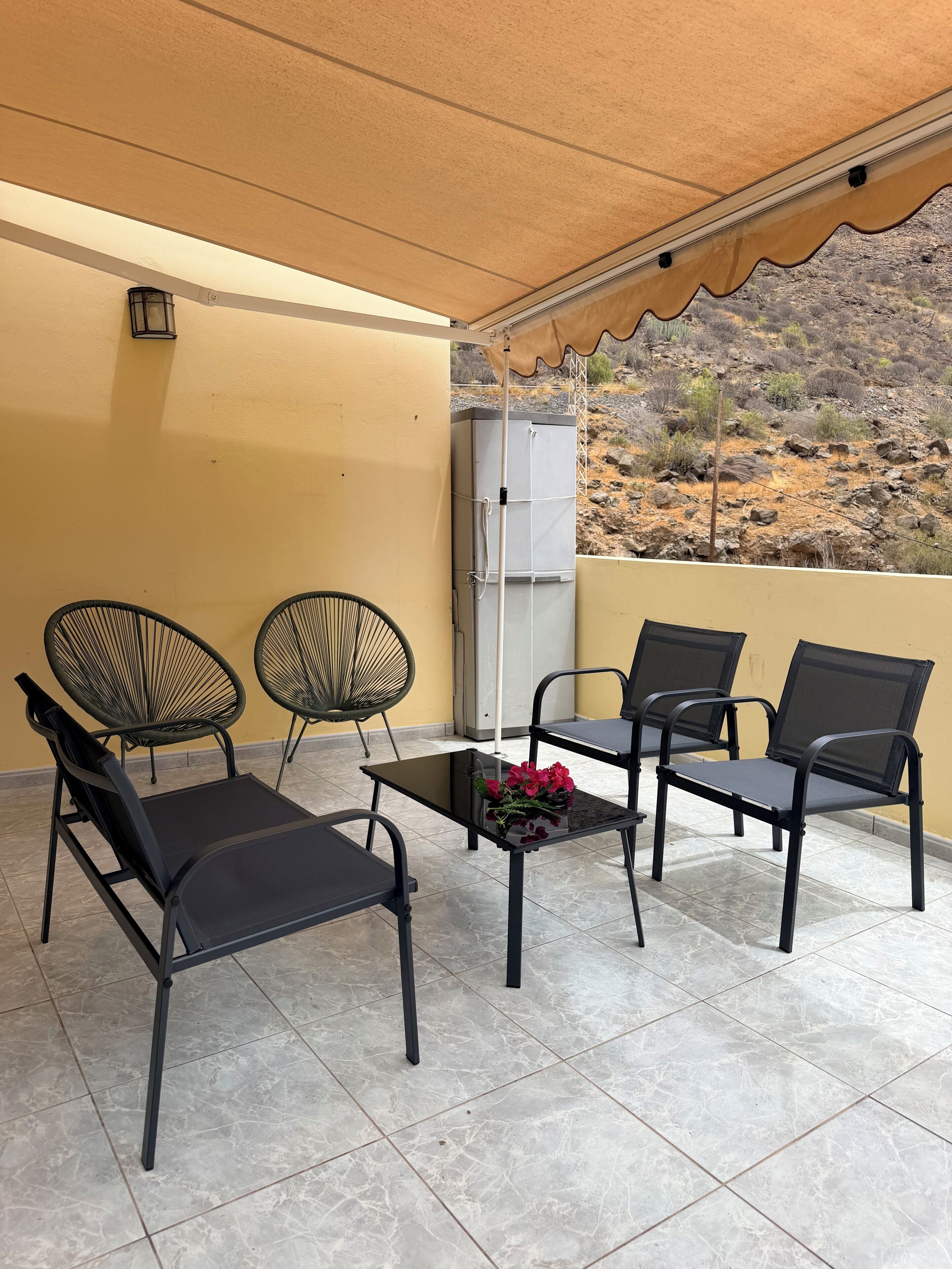 A covered outdoor seating area with two chairs and a table, a refrigerator in the background, and a view of a rocky landscape.